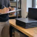 Hands recording an equipment inventory beside retired laptops and desktop computers